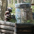 Extreme team sport. The player in the paintball looks out of hiding. Camouflage suit, protective mask and weapons. Tires, barrels, Royalty Free Stock Photo