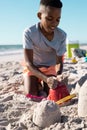 Playful african american boy building a sandcastle with pail on beach against clear sky, copy space