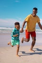 Playful african american boy holding young father\'s hands and running at beach against blue sky Royalty Free Stock Photo