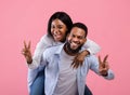 Playful black guy giving his girlfriend piggyback ride, showing peace gesture on pink studio backgroud Royalty Free Stock Photo