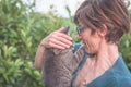 Playful domestic cat held and cuddled by smiling woman with eyeglasses. Outdoor setting in green home garden. Shallow depth of fie