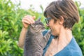 Playful domestic cat held and cuddled by smiling woman with eyeglasses. Outdoor setting in green home garden. Shallow depth of fie