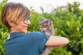 Playful domestic cat held and cuddled by smiling woman with eyeglasses. Outdoor setting in green home garden. Shallow depth of fie