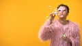 Playful female teenager blowing soap bubbles on yellow background, youth party