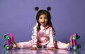 Playful kid girl with nice buns in grey hooded jumpsuit costume and funny roller skates sits with her legs apart and show tongue