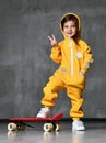 Playful kid girl in yellow hooded jumpsuit and white sneakers stands on skateboard, show peace sign and keep her hand in pocket