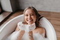 Playful lady having fun while lying in bathtub full of foam, blowing soap bubbles and smiling, copy space