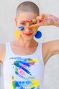 Playful portrait of a young gorgeous female artist painter covered in paint, looking and smiling at camera through her fingers.