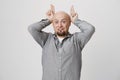 Playful positive bald male with beard has cheerful expression, wears gray shirt, behaves as rabbit, holds fingers above