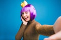 Playful stylish selfie portrait brightful young woman with cut purple hair, golden crown showing tongue on blue Royalty Free Stock Photo