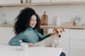 Playful woman with Afro haircut, pets her breed dog, have fun together, pose in cozy kitchen, drink coffee, laugh happily. Young