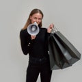 Playful young caucasian woman dressed in black smiling at camera and holding megaphone, posing with shopping bags Royalty Free Stock Photo
