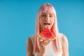 Playful young woman with pink hair holding a slice of watermelon, posing with mouth open isolated over blue studio Royalty Free Stock Photo