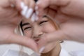 A playful young woman posing closely to the camera and framing her face with a hand heart gesture. Isolated on a white background