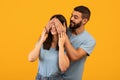 Playing hide and seek. Happy arab bearded guy covering his pretty girlfriend eyes from back, yellow background Royalty Free Stock Photo