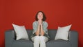 Pleading young woman keep palms together implore beg for favor or help sitting on sofa. Red background