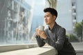 Pleasant asian man enjoying his morning coffee while standing near office window overlooking cityscape Royalty Free Stock Photo