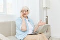 a pleasant, happy grandmother is sitting on the couch talking on the phone with her loved ones and holding a laptop on Royalty Free Stock Photo
