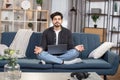 Pleasant likable young arabian indian man, sitting in lotus pose with laptop pc on sofa at living room, and meditating