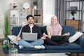 Pleasant likable young arabian muslim man and woman in hijab, sitting in lotus pose with laptop pc on sofa at living