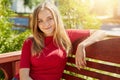 Pleasant-looking blonde female wearing casual red sweater sitting at comfortable wooden bench against green park background having