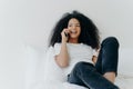 Pleasant looking curly woman has pleasant conversation, talks with friend via smartphone while relaxes in bed, has joyful