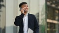 Pleasant millennial male entrepreneur in black suit having phone conversation while standing in front of business center