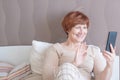 Pleasant senior woman with short red hair in home clothes is sitting on the bed with a smartphone and communicates with friends or