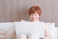 Pleasant senior woman with short red hair is sitting on the bed with a laptop, looking at the monitor and smiling