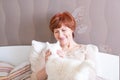 Pleasant senior woman with short red hair is sitting on the bed and petting a cute white cat