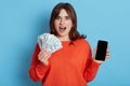 Pleasantly shocked young woman in orange sweater holding mobile phone with blank screen and fan of money, girl with cash isolated