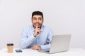 Please, be quiet! Funny positive man employee sitting office workplace with laptop, asking for silence or secrecy