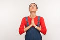 Please god, help! Portrait of imploring worried girl with hair bun in denim overalls looking up and begging, praying Royalty Free Stock Photo