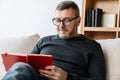 Pleased adult unshaven man writing down notes while sitting on couch