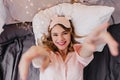 Pleased blonde girl lying on dark sheet. Indoor overhead photo of smiling cheerful lady in pink eyemask posing in bed..