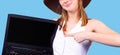 Pleased blonde woman in short white top and hat showing blank laptop computer screen pointing on it with index finger. Royalty Free Stock Photo