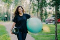 Pleased brunette woman dressed in black t shirt, holds rolled up karemat and fitness ball, prepares for aerobic exercises, smiles