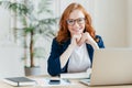 Pleased cheerful red haird female economist develops financial startup project, poses in office interior, works in business sphere Royalty Free Stock Photo