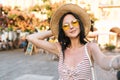 Pleased cute girl wearing trendy yellow sunglasses spending time near outdoor restaurant waiting friends and making Royalty Free Stock Photo