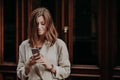 Pleased delighted female youngster watches video on smart phone, reads banking notification, dressed in white raincoat, poses agai