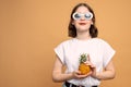 Pleased european girl in white sunglasses holds pineapple in her hands and looking at the camera, orange wall on the