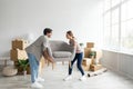 Pleased european young male and female carry armchair in empty room with cardboard boxes with belongings