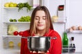 Pleased female holds saucepan, smells pleasant oudour, cooked delicious soup with vegetables, stands at kitchen near opened fridge