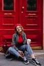 Pleased girl in glasses and black gumshoes drinks coffee, sitting near red door. Stunning young woman in beret posing on