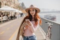 Pleased girl with sly smile holding phone near street river. Outdoor portrait of wonderful ginger woman in casual outfit