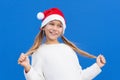 Pleased girl in white sweater and Santa hat holding pigtails smiling away isolated on blue background Royalty Free Stock Photo