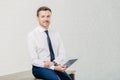 Pleased male entrepreneur makes payment online on digital tablet, dressed in elegant white shirt, tie and trousers, sits on wooden