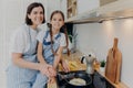 Pleased mother with little helping daughter near cooker, prepare breakfast for whole family, fry eggs on pan, pose in modern