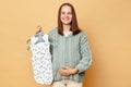 Pleased positive happy young pregnant woman with baby bodysuits isolated over beige background touching her belly looking at Royalty Free Stock Photo