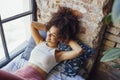 Pleased relaxed African American ethnicity girl in homewear sitting on windowsill